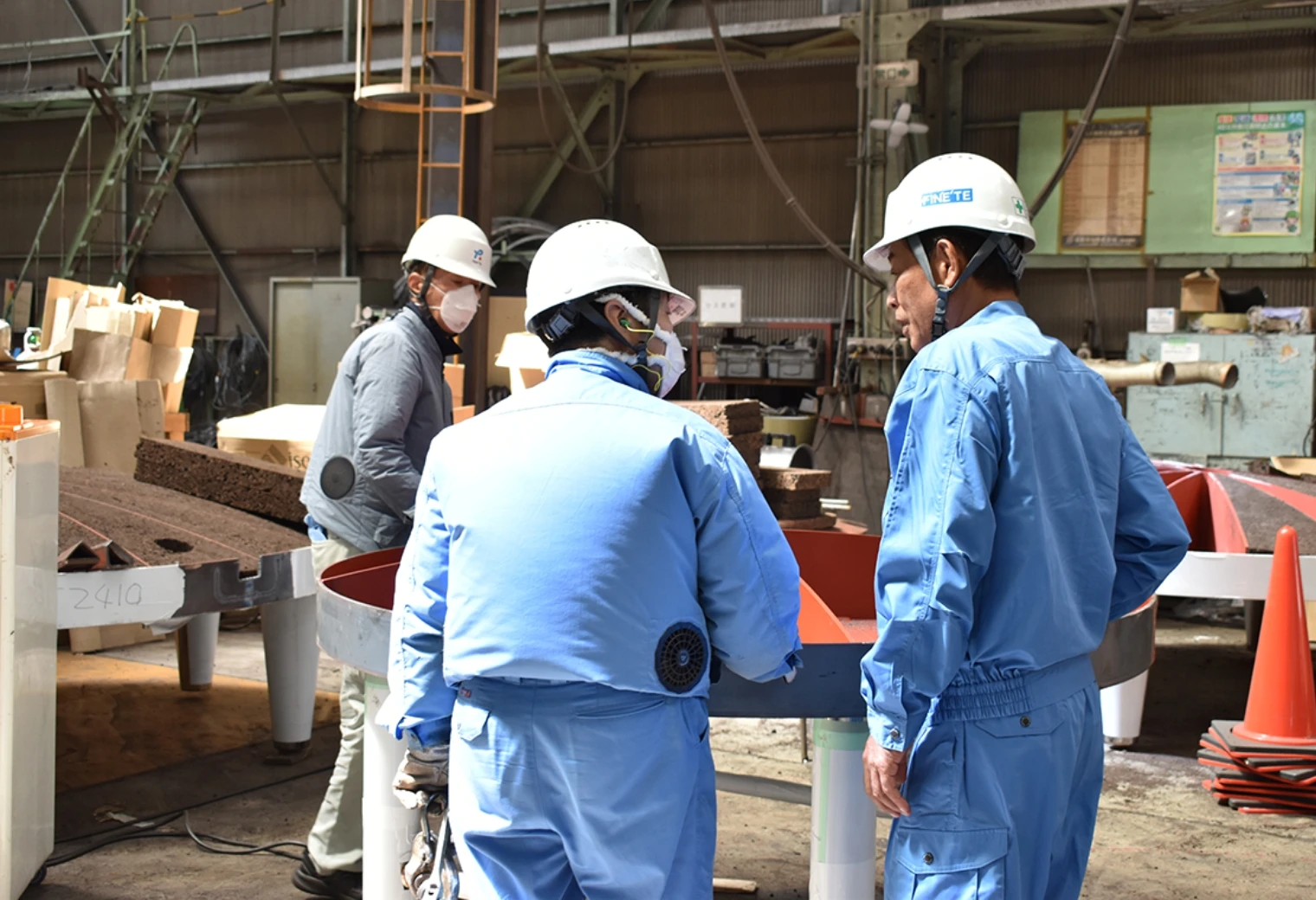 工場内で製品を囲みながら打ち合わせを行う、ヘルメット姿の技術者と作業員たちの様子