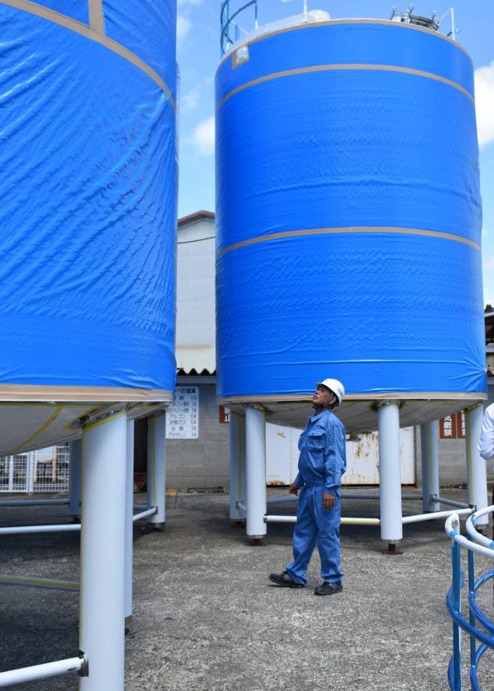 大型の青いタンク設備の前で、作業服とヘルメットを着用した作業員が点検している様子