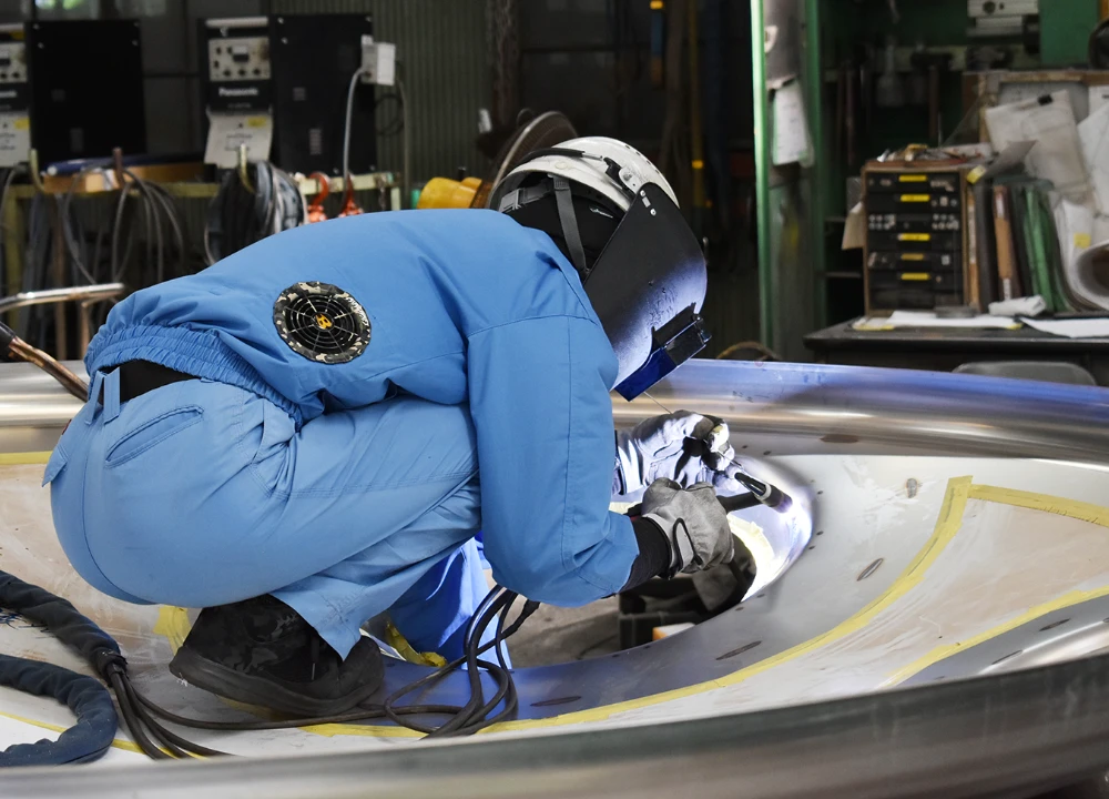 工場で青い作業服を着た技術者が、大きな金属部品の溶接作業に集中している様子