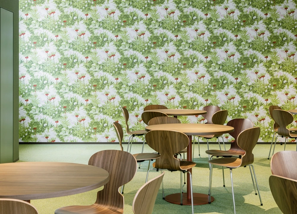 植物柄の壁紙がおしゃれな、木製の丸テーブルと椅子が並ぶ明るい休憩スペース