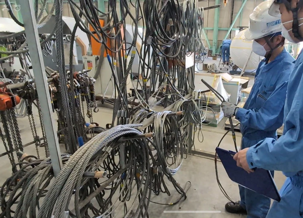 工場の資材置き場で、多くのワイヤーロープを点検し、クリップボードで管理を行う二人の作業員