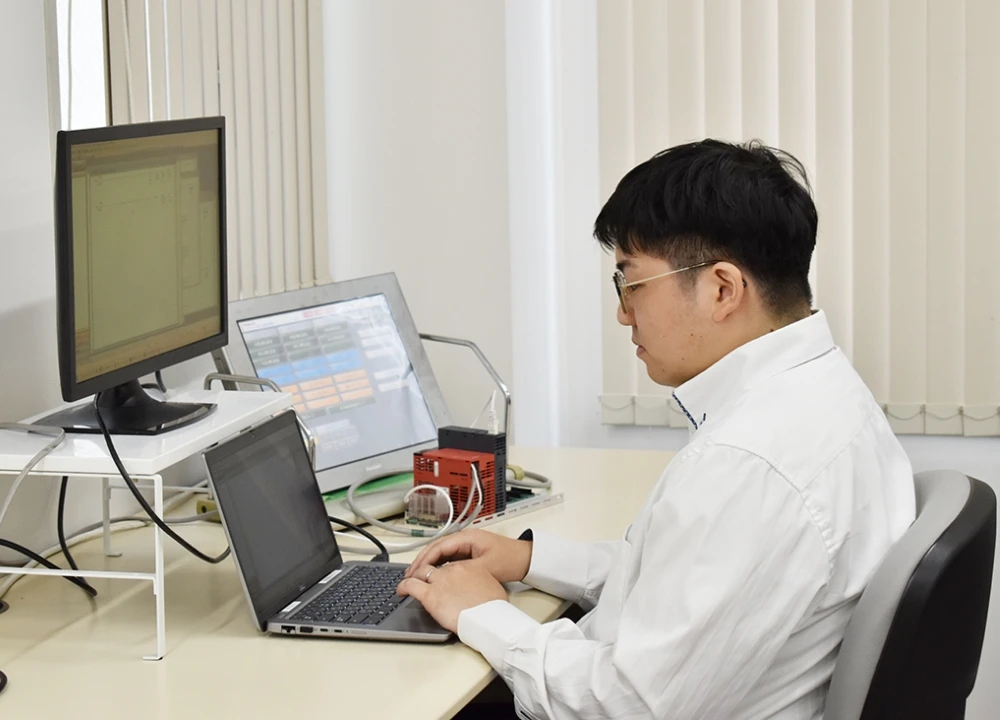 デスクでノートパソコンを操作し、複数のモニターや制御機器を使いながら設計やプログラミングを行う技術者