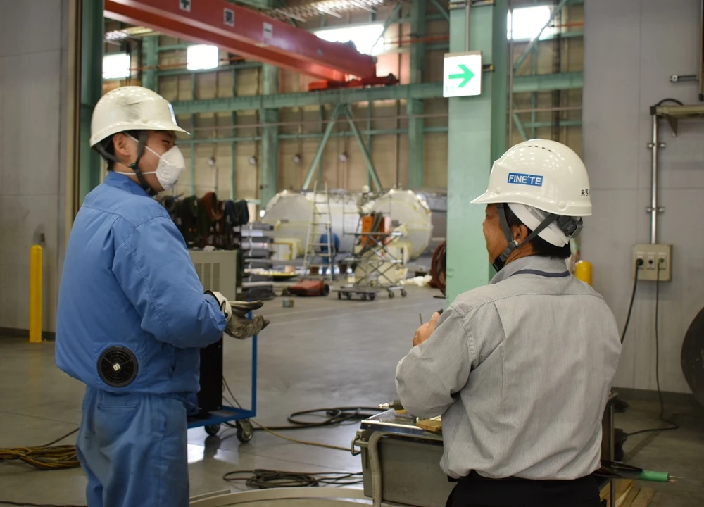 ヘルメットを着用した二人のスタッフが、工場の広い作業現場で進捗状況について話し合っている様子