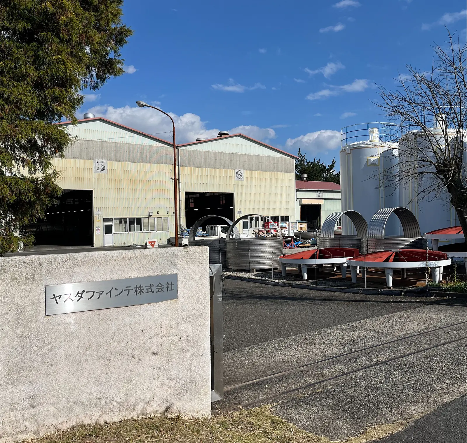 工業用タンクと工場の外観、青空の下のヤスダファインテ株式会社の入口。